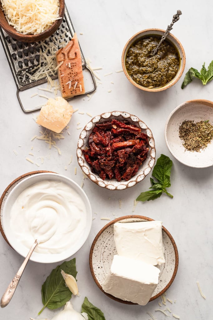 Sun-dried tomatoes and pesto in bowls next to cheese ingredients.
