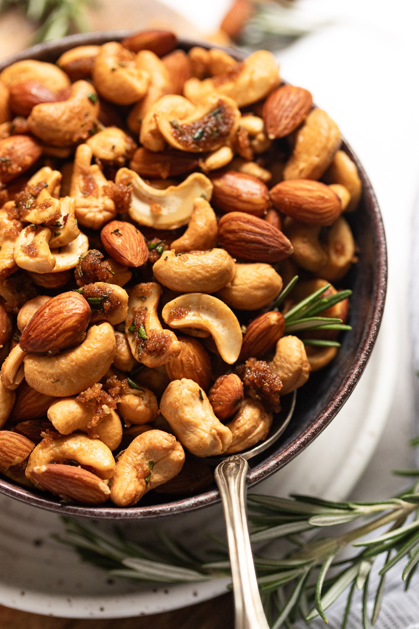 Side view of bowl of roasted sweet and spicy nuts with spoon and rosemary garnish.