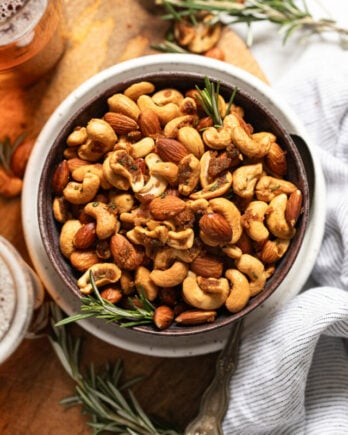 Bowl of roasted rosemary nuts on plate next to drinks and linen.