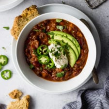 spicy vegetarian bean chili with avocado, jalapeno, and sour cream in white bowl with bread by fork in the kitchen