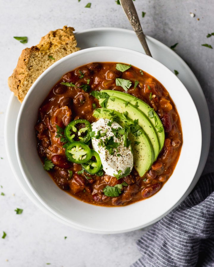 spicy vegetarian bean chili with avocado, jalapeno, and sour cream in white bowl with bread by fork in the kitchen