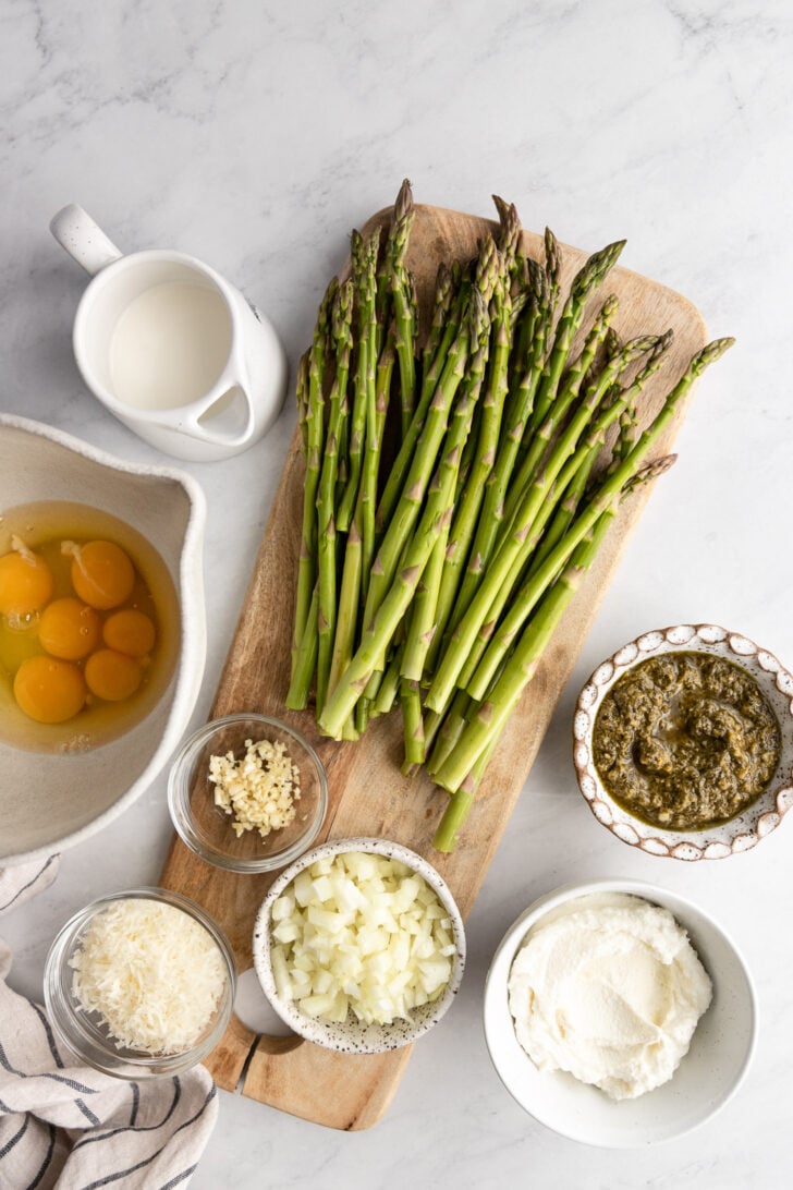 Asparagus spears on cutting board next to bowls of onion, ricotta, pesto, parmesan, garlic, eggs, and half and half.