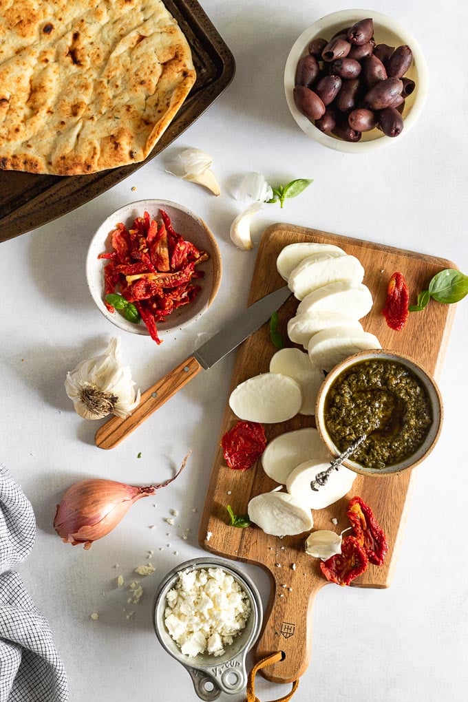 Cheese and pesto on cutting board next to sun-dried tomatoes, olives, shallot, and naan flatbread.
