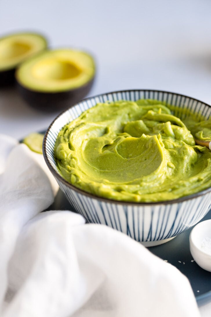 Side view of avocado sauce in bowl with white linen and avocado halves in background.