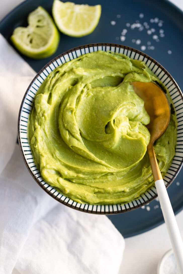 Bowl of avocado sauce on blue plate with limes and spoon.