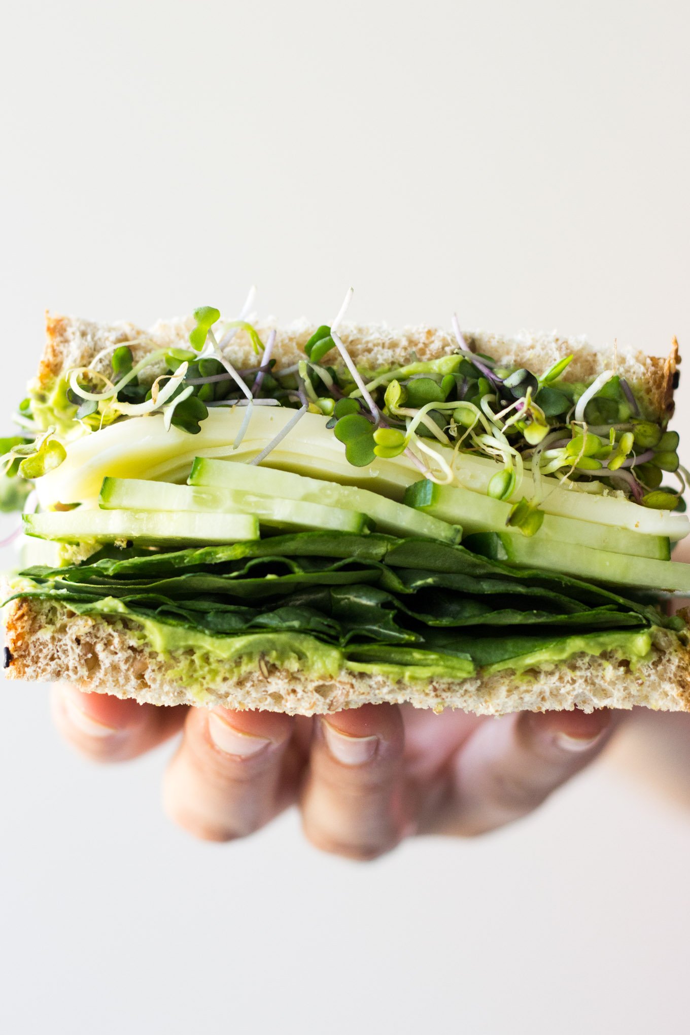 Hand holding vegetarian sandwich with layers of microgreens, cucumber, cheese, spinach, and avocado.