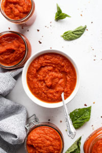 Bowl of tomato sauce with spoon surrounded by other jars.