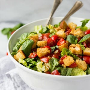 White bowl with bruschetta salad, croutons, and serving utensils.