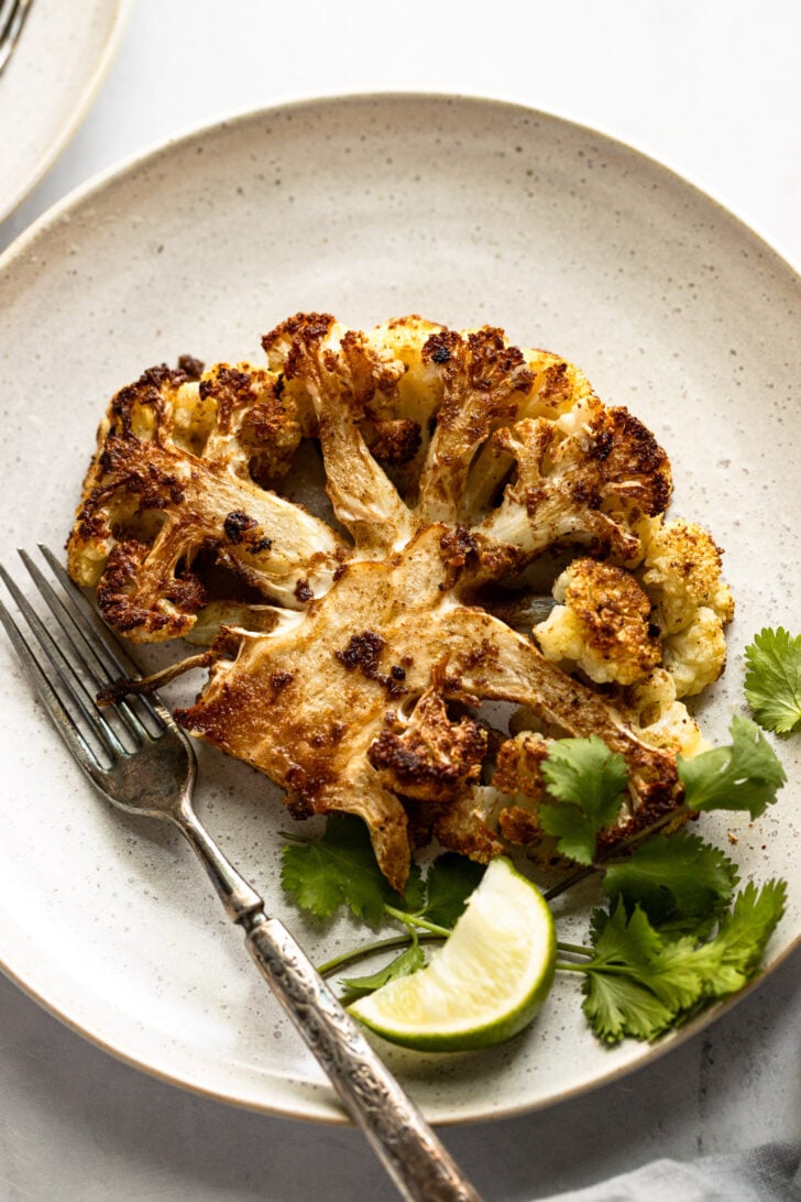 Cauliflower steak on plate next to lime and cilantro.