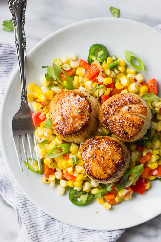 White plate with corn and red peppers topped with scallops.