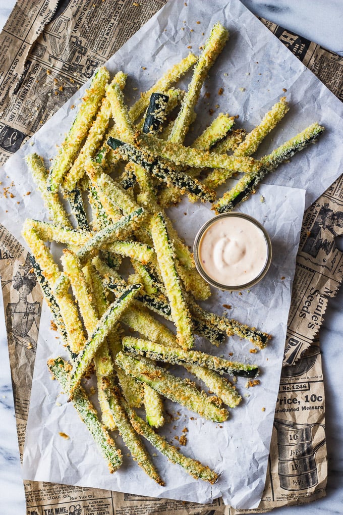 Crispy zucchini fries on parchment paper and old newspaper with aioli next to them.