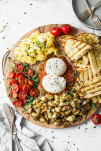 round wood board filled with burrata, tomatoes, potatoes, toast, and eggs alongside silverware