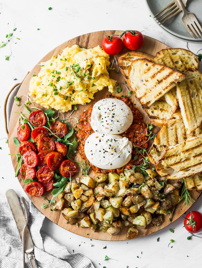round wood board filled with burrata, tomatoes, potatoes, toast, and eggs alongside silverware