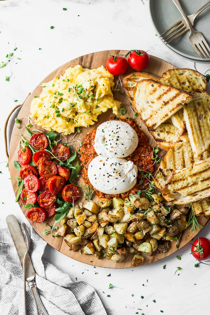 Round wood board filled with burrata, tomatoes, potatoes, toast, and eggs alongside silverware.