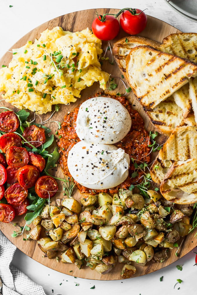 Close up round wooden breakfast board with burrata, tomatoes, eggs, toast, potatoes.