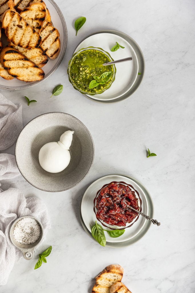 Burrata, pesto, and tomato jam in separate bowls next to grilled baguette.