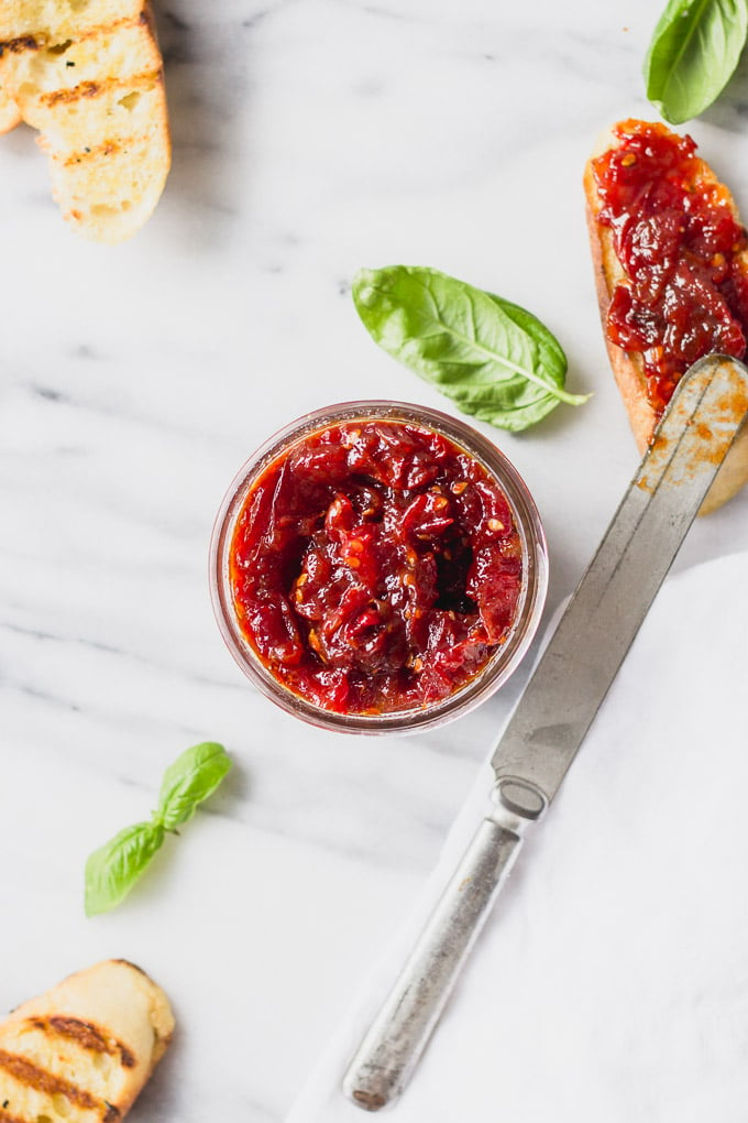 Overhead mason jar with tomato jam next to knife spreading on baguette.