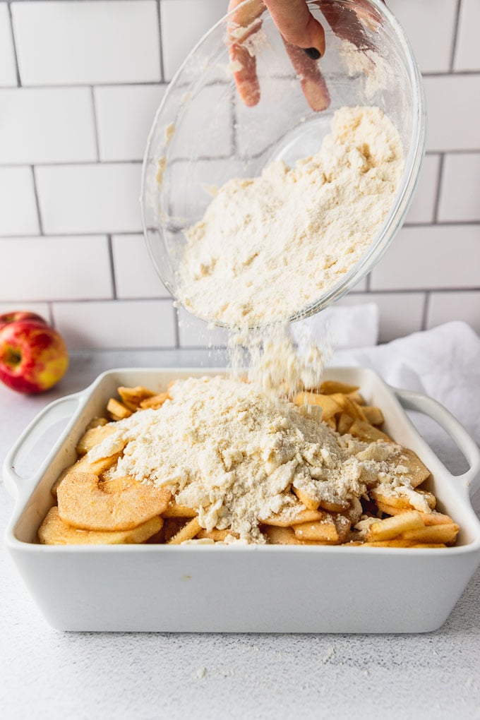Pouring apple crisp topping onto sliced apples in baking dish.
