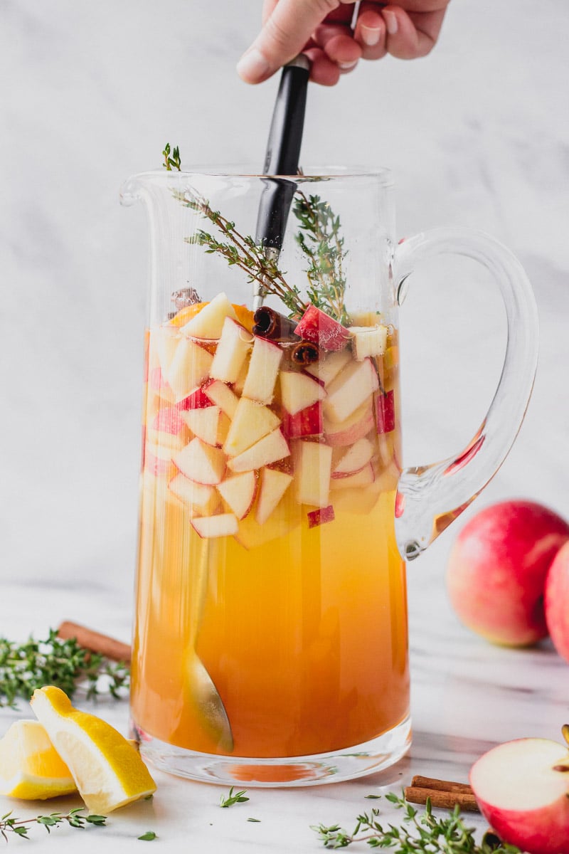 stirring apple cider sangria in pitcher with cinnamon, thyme, and apples by fork in the kitchen