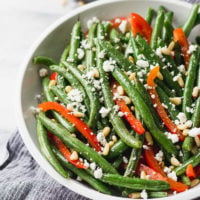 green beans with red bell pepper, pine nuts, and goat cheese in white bowl by fork in the kitchen