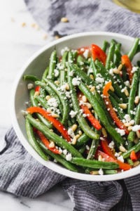 green beans with red bell pepper, pine nuts, and goat cheese in white bowl by fork in the kitchen