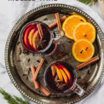 glass mug with mulled red wine, cinnamon, and orange slices on tray by fork in the kitchen