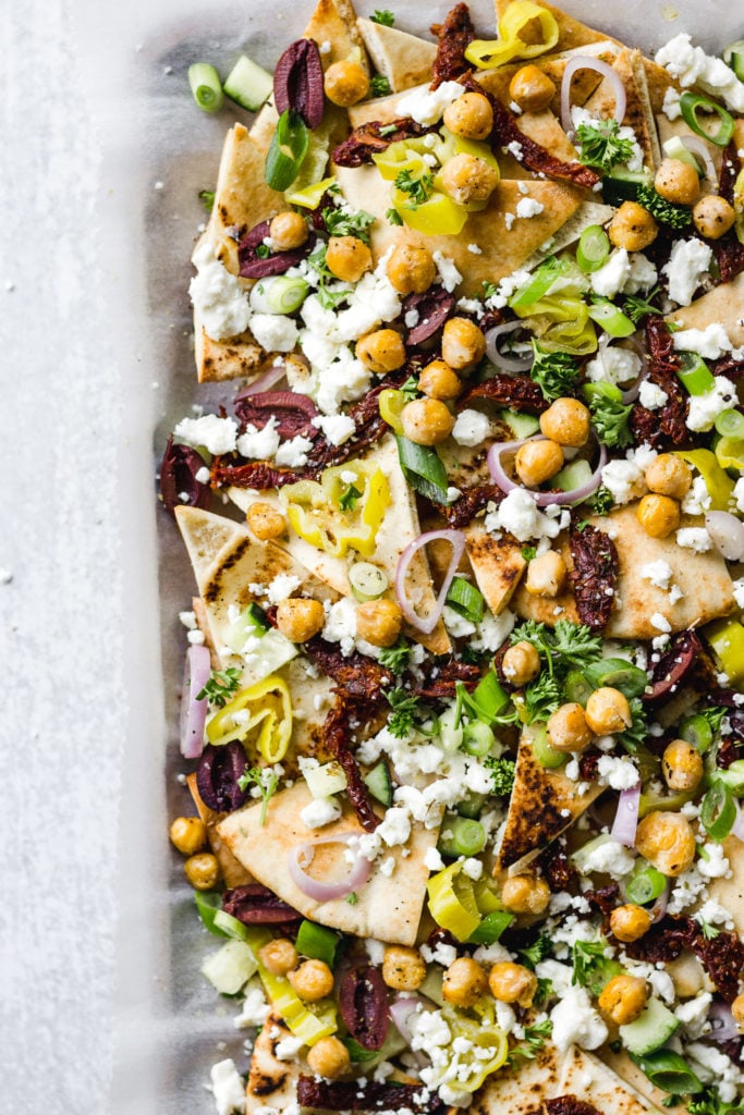 Baking sheet with pita bread triangles topped with shallot, banana peppers, chickpeas, sun-dried tomatoes, and olives.