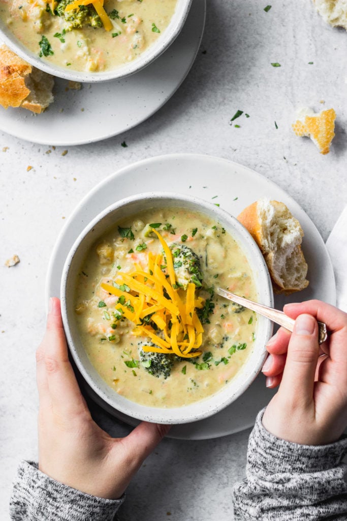 sopa de queijo cheddar de couve-flor de brócolos em tigela em cima do prato a ser segurado por uma mão e outra colher de agarrar à mão