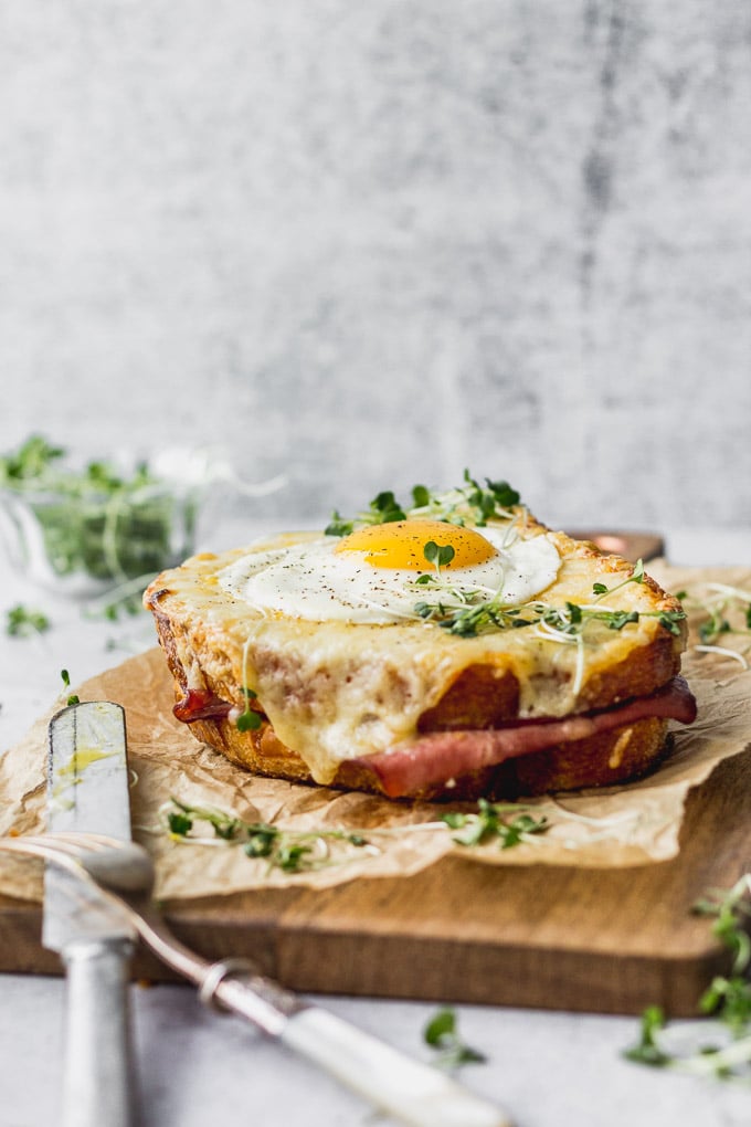 Croque madame sandwich on cutting board with microgreens next to fork and knife.