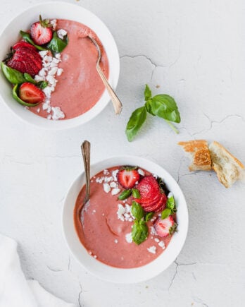 two white bowls with pink strawberry basil gazpacho next to baguette and white linen