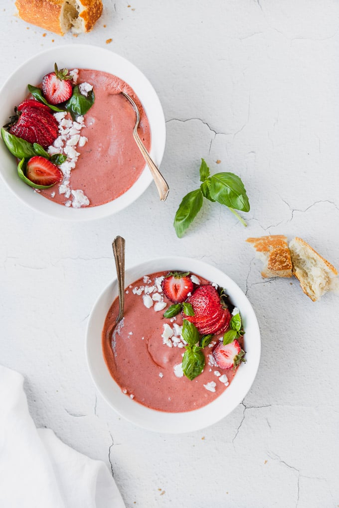 two white bowls with pink strawberry basil gazpacho next to baguette and white linen
