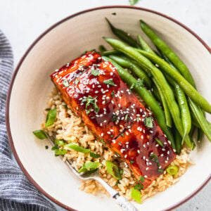 Bowl with rice, green onions, teriyaki salmon, and fresh green beans next to blue linen.