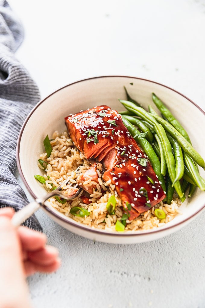 hand with fork taking a bite of teriyaki salmon in bowl