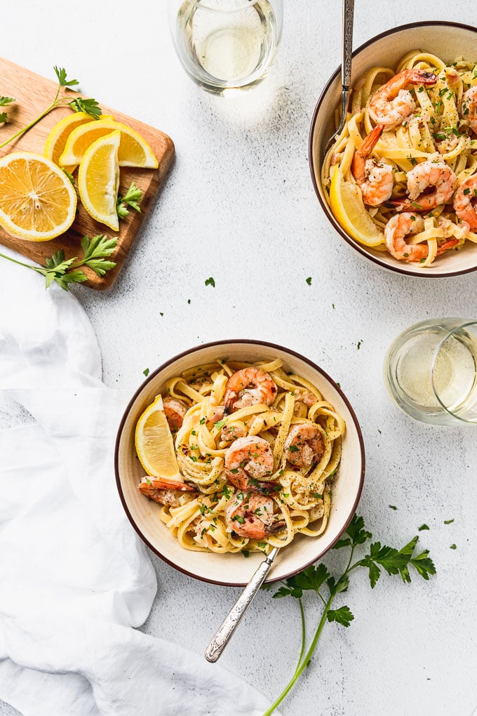 Two bowls of pasta with shrimp and lemon wedges next to wine glasses.