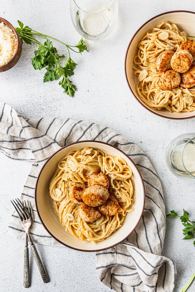 Two bowls of creamy garlic pasta topped with seared scallops next to parmesan bowl and wine glass.