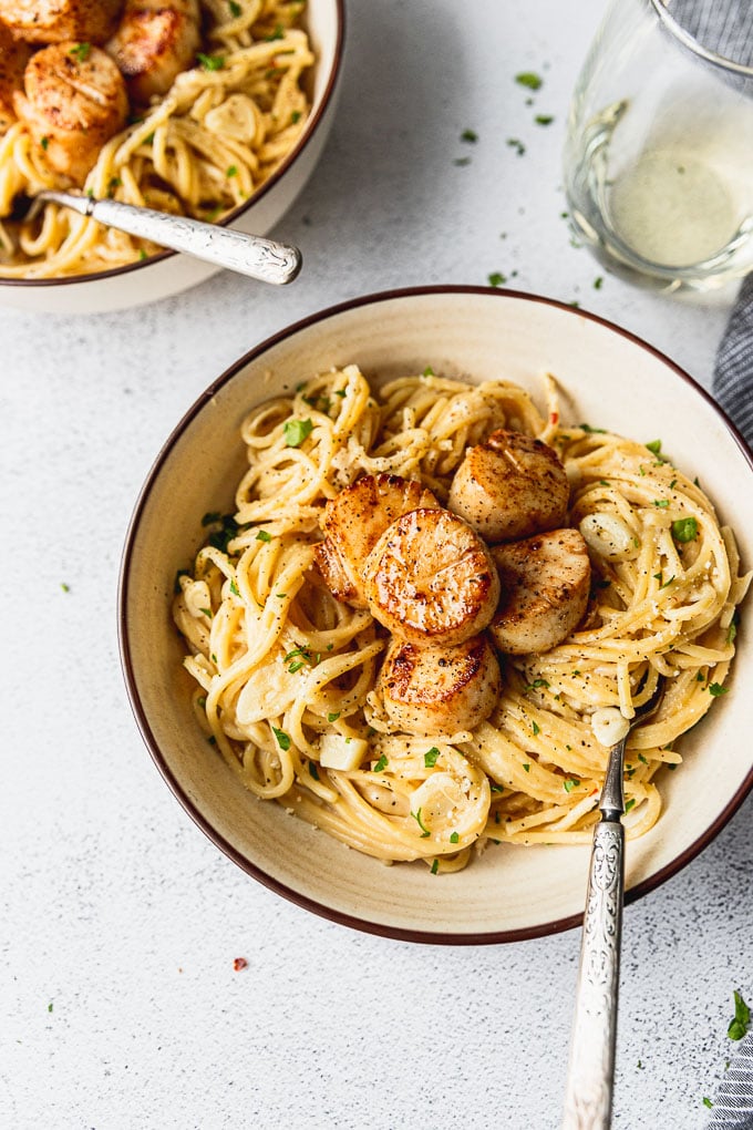 Up close look into a cream colored bowl with spaghetti pasta topped with seared scallops and a fork in the bowl.