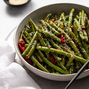 Bowl of spicy green beans next to white linen and sesame seeds.