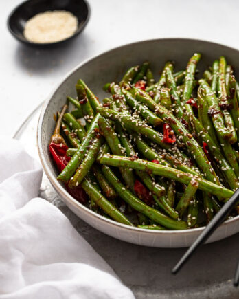 Bowl of spicy green beans next to white linen and sesame seeds.