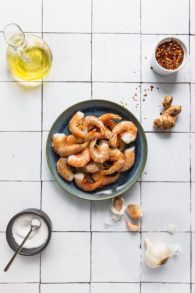 plate of raw shrimp next to ginger root, red pepper flakes, garlic cloves, salt, and olive oil for firecracker shrimp ingredients