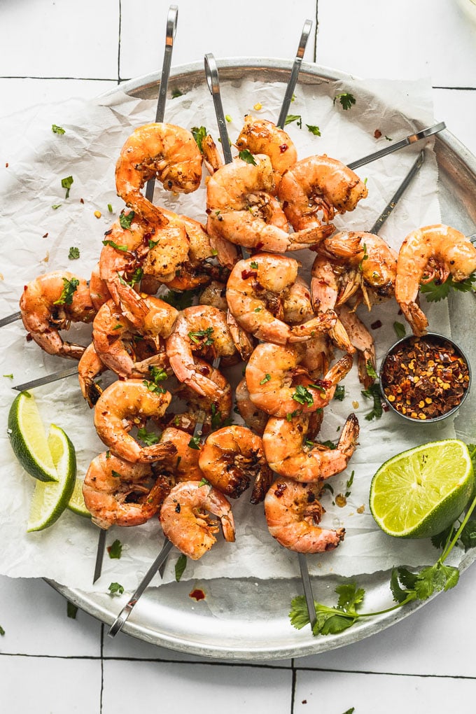 overhead view of skewers on top of each other on a tray in diagonal directions with shrimp next to lime wedges and cup of red pepper flakes