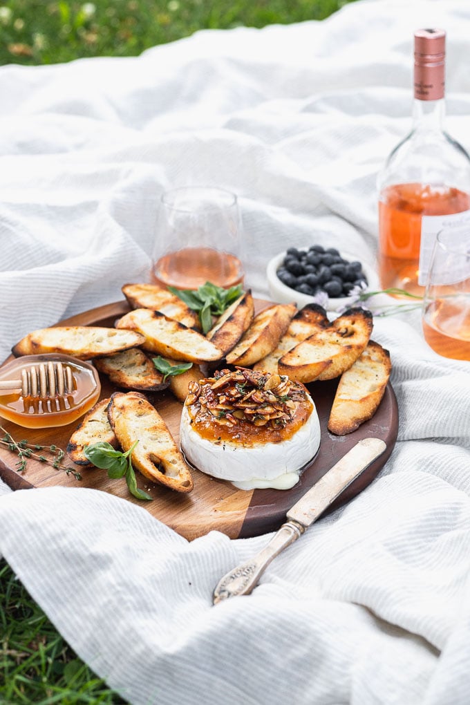 wood serving tray on picnic blanket with wine, grilled brie and baguette slices, and honey