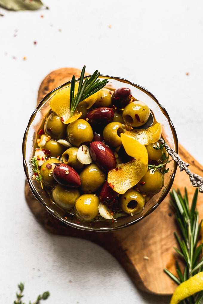 glass bowl filled with marinated olives with a spoon on top of a wood serving tray