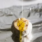 raw garlic bulb with top cut off on foil and a drizzle of oil being poured