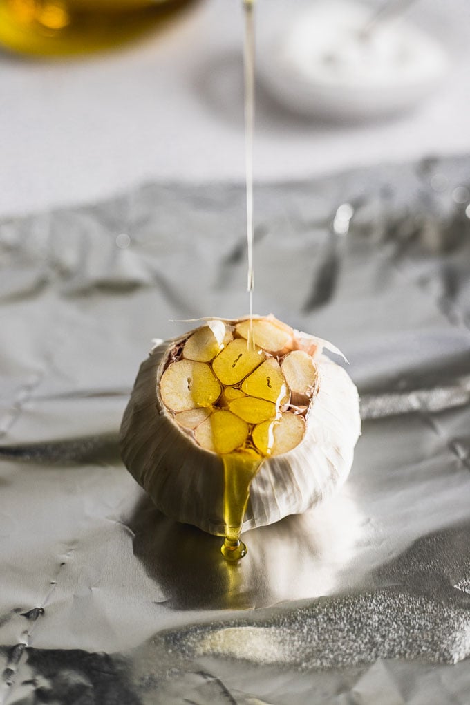 raw garlic bulb with top cut off on foil and a drizzle of oil being poured