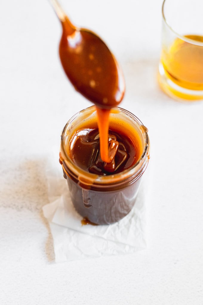 bourbon caramel sauce in jar with spoon drizzling next to a glass of bourbon