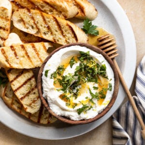 A large white plate topped with grilled baguette slices and a wooden bowl filled with ricotta next to two wine glasses.
