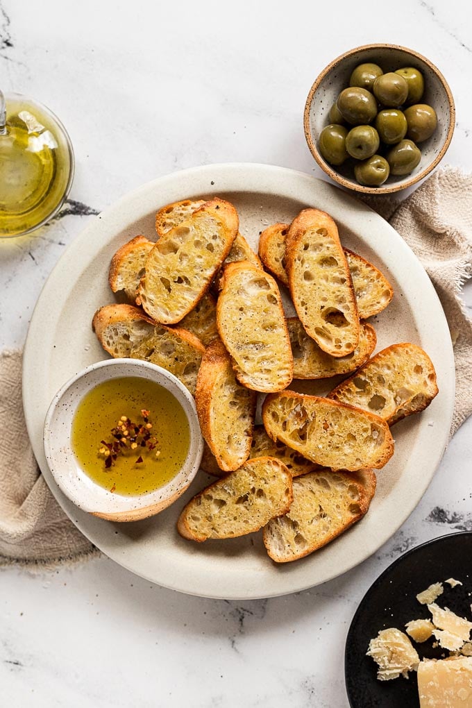 Plate of crostini with small bowl of olive oil next to a bowl of olives and parmesan cheese.