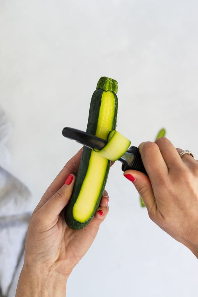Hands holding zucchini with a vegetable peeler peeling a ribbon.