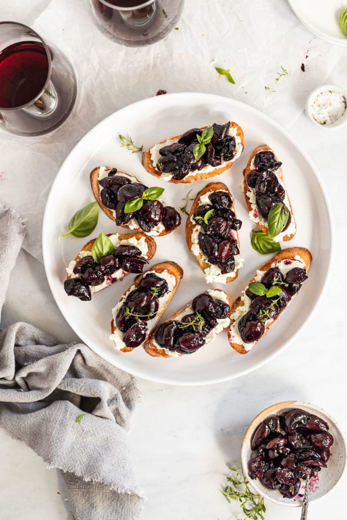 Roasted cherry crostini on white plate next to red wine glasses.