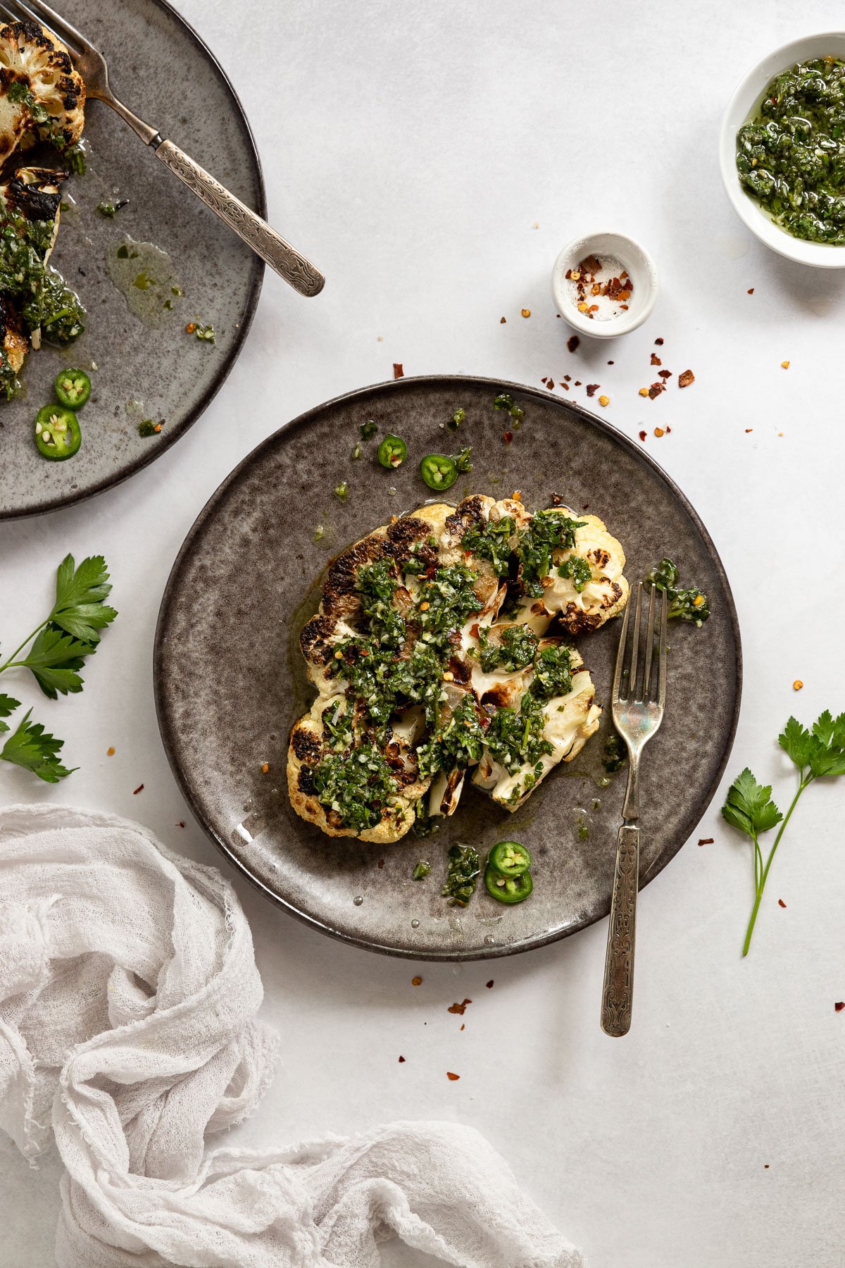 Cauliflower steaks on plates with forks next to linen and chimichurri sauce.